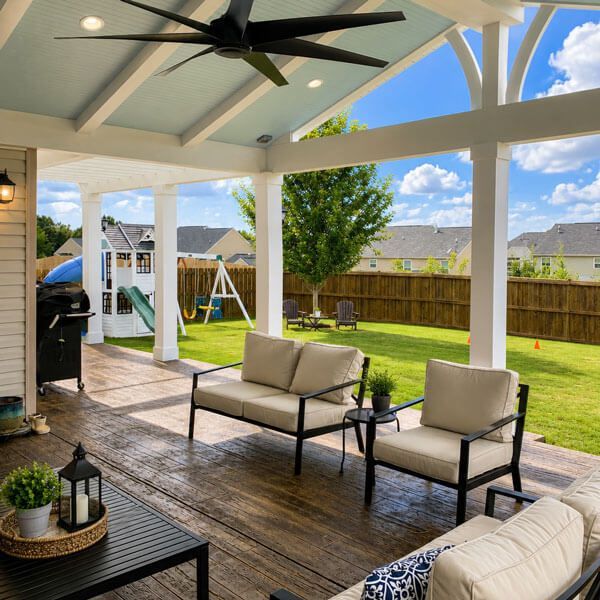 Covered patio with seating, ceiling fan, and grill overlooking a sunny backyard and swing set.