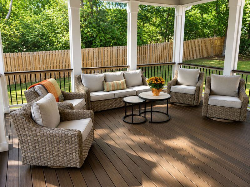 Covered deck with wicker patio furniture, beige cushions, round table, and a wooden fence in the background.