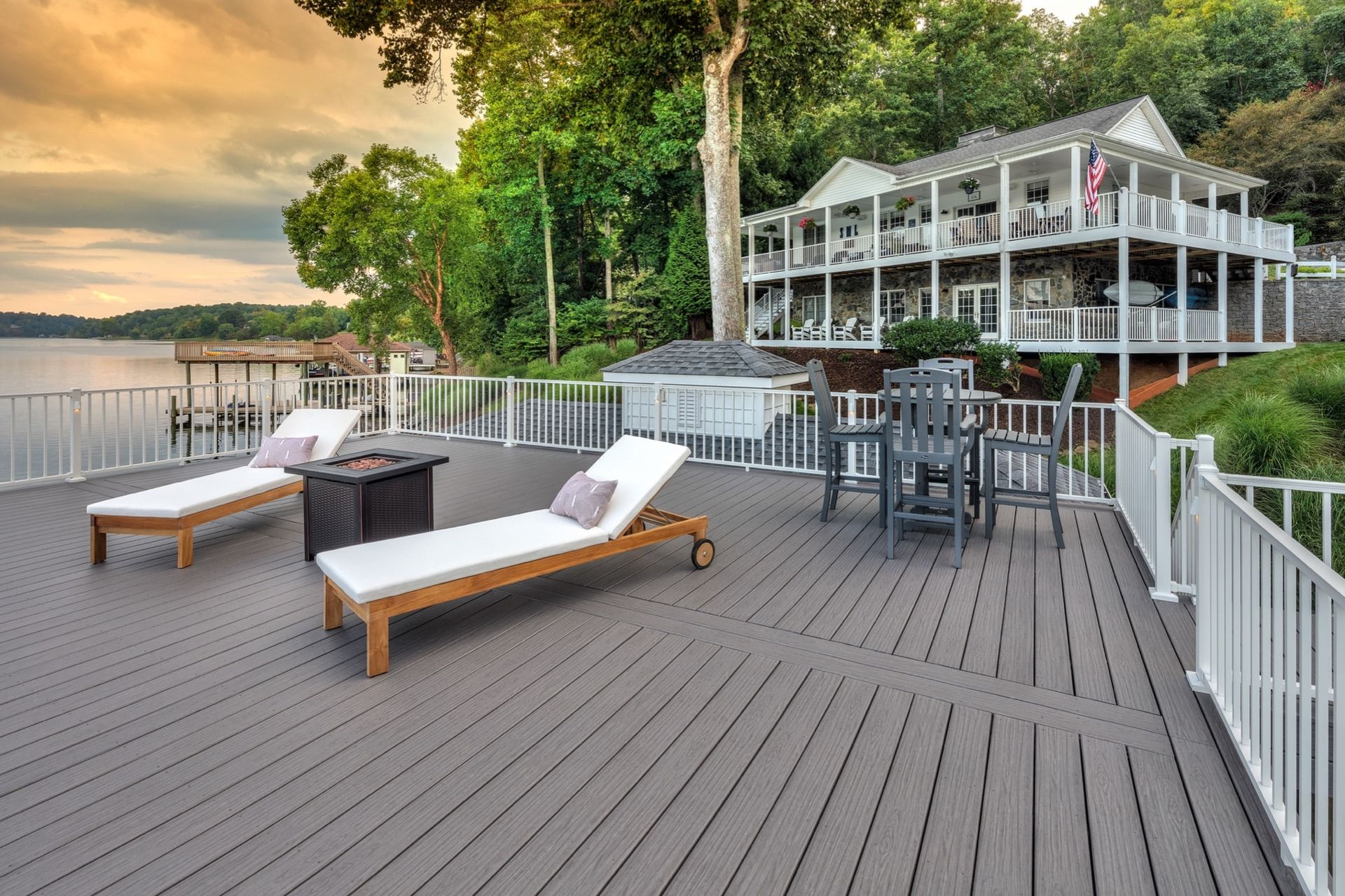 Lakeside deck with lounge chairs, dining tables, and a white house at sunset