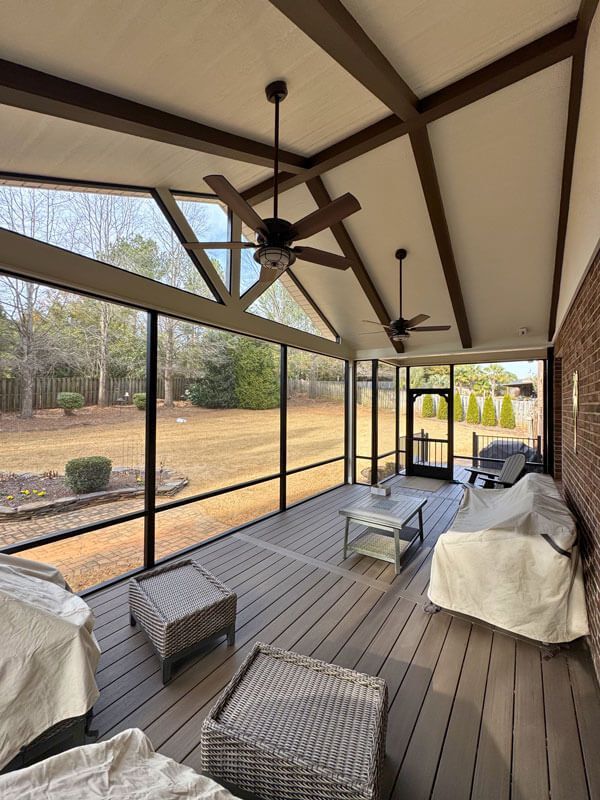 Screened porch with ceiling fans, wicker seating, and a covered sofa overlooking a wooded yard.