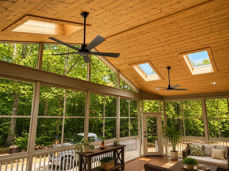 Bright sunroom with wood ceiling, skylights, ceiling fans, and large windows overlooking a green forest.