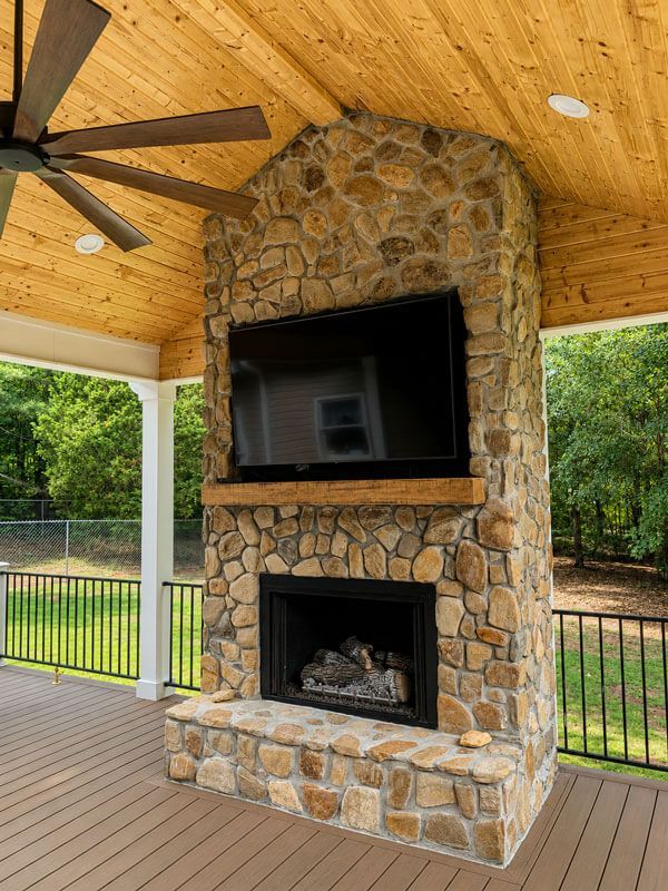 Covered patio with stone fireplace and mounted TV under a wood stainedceiling with a ceiling fan
