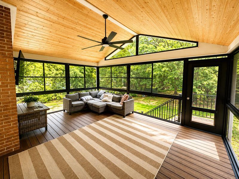 Spacious screened porch with wood ceiling, ceiling fan, gray sectional, striped rug, and forest view