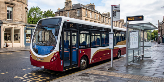 Red and blue city bus stopped at a curbside stop beside a glass shelter and station sign