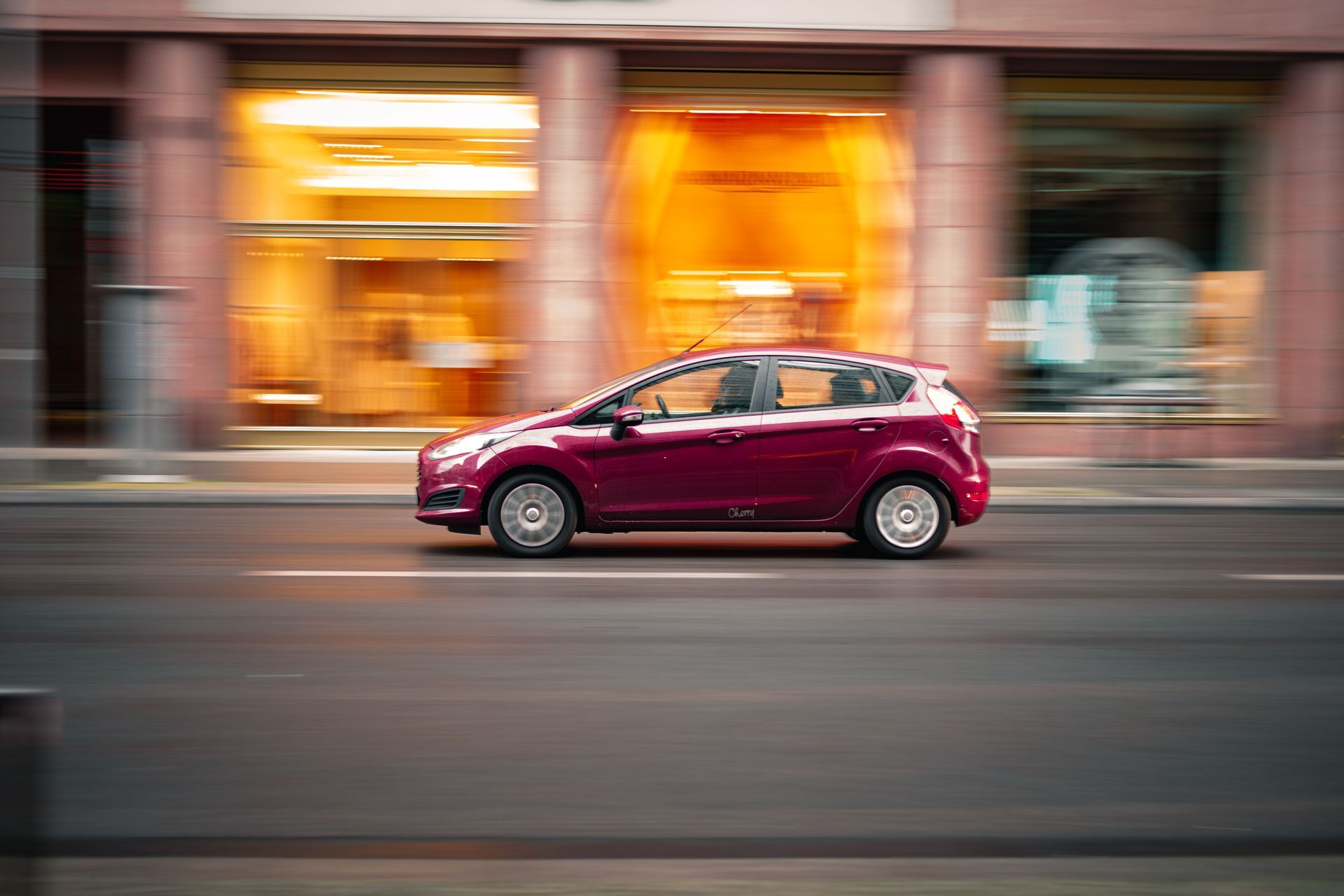 Red hatchback driving past storefronts at dusk, blurred by motion