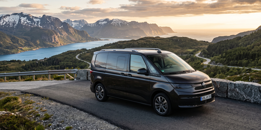 Black van parked on a mountain road overlooking a lake and snowy peaks at sunset