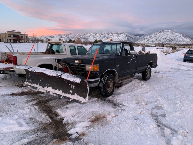 Black pickup truck with plow in snowy lot at sunset, mountains in the background
