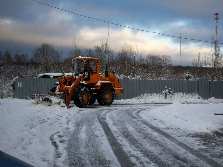 Orange front loader clearing snow in a snowy yard beside a fence under cloudy skies