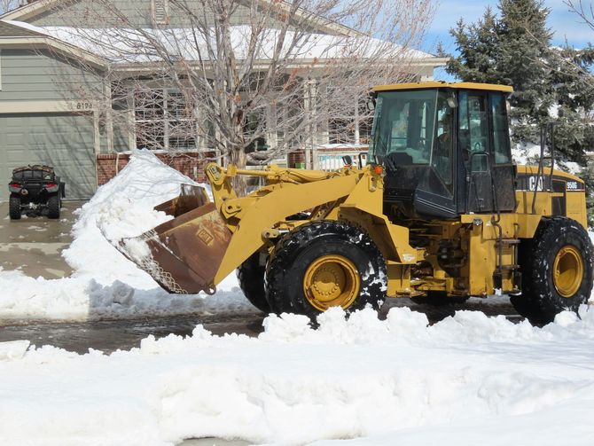 Yellow front loader pushing snow in a residential driveway with houses in the background