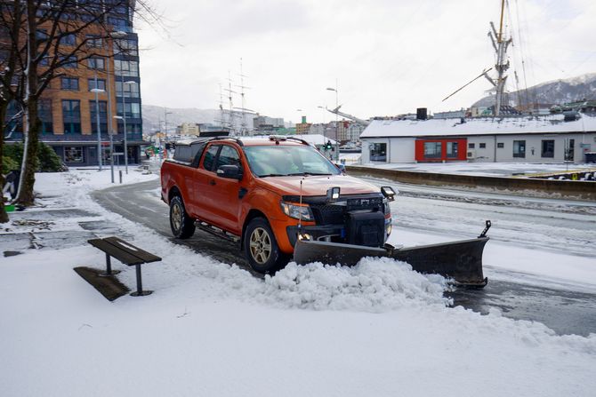 Orange pickup truck with snow plow clearing a snowy street beside waterfront buildings