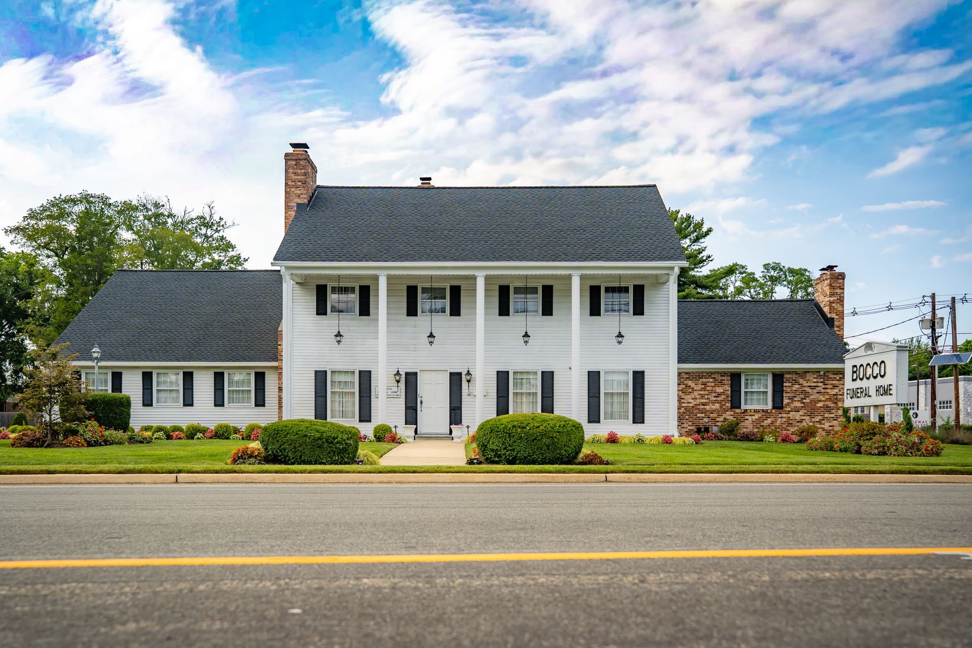 White colonial-style house with gray roof and brick chimneys beside a road under a partly cloudy sky