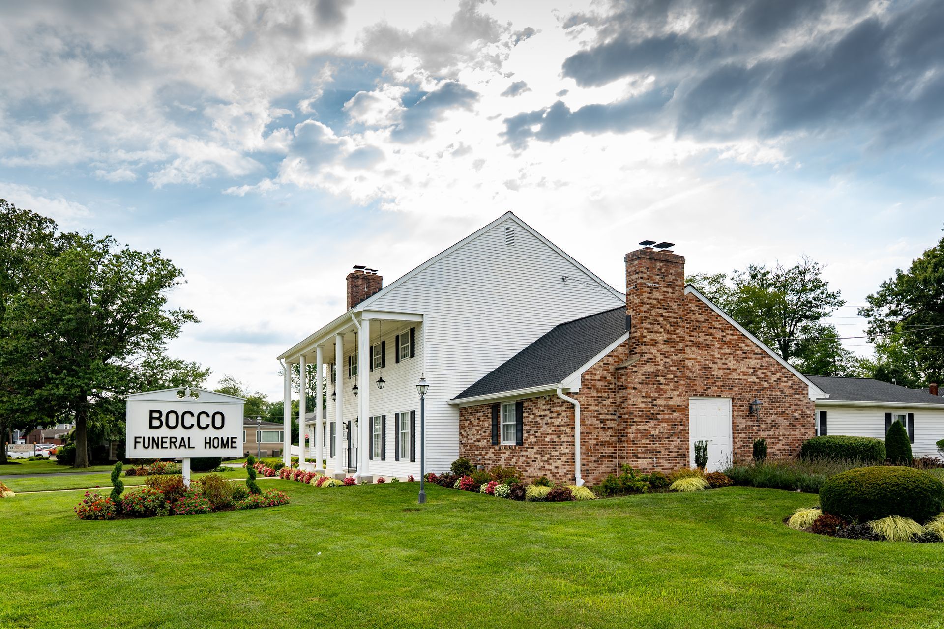 Bocco Funeral Home building with white siding, brick chimneys, and a green lawn under a cloudy sky