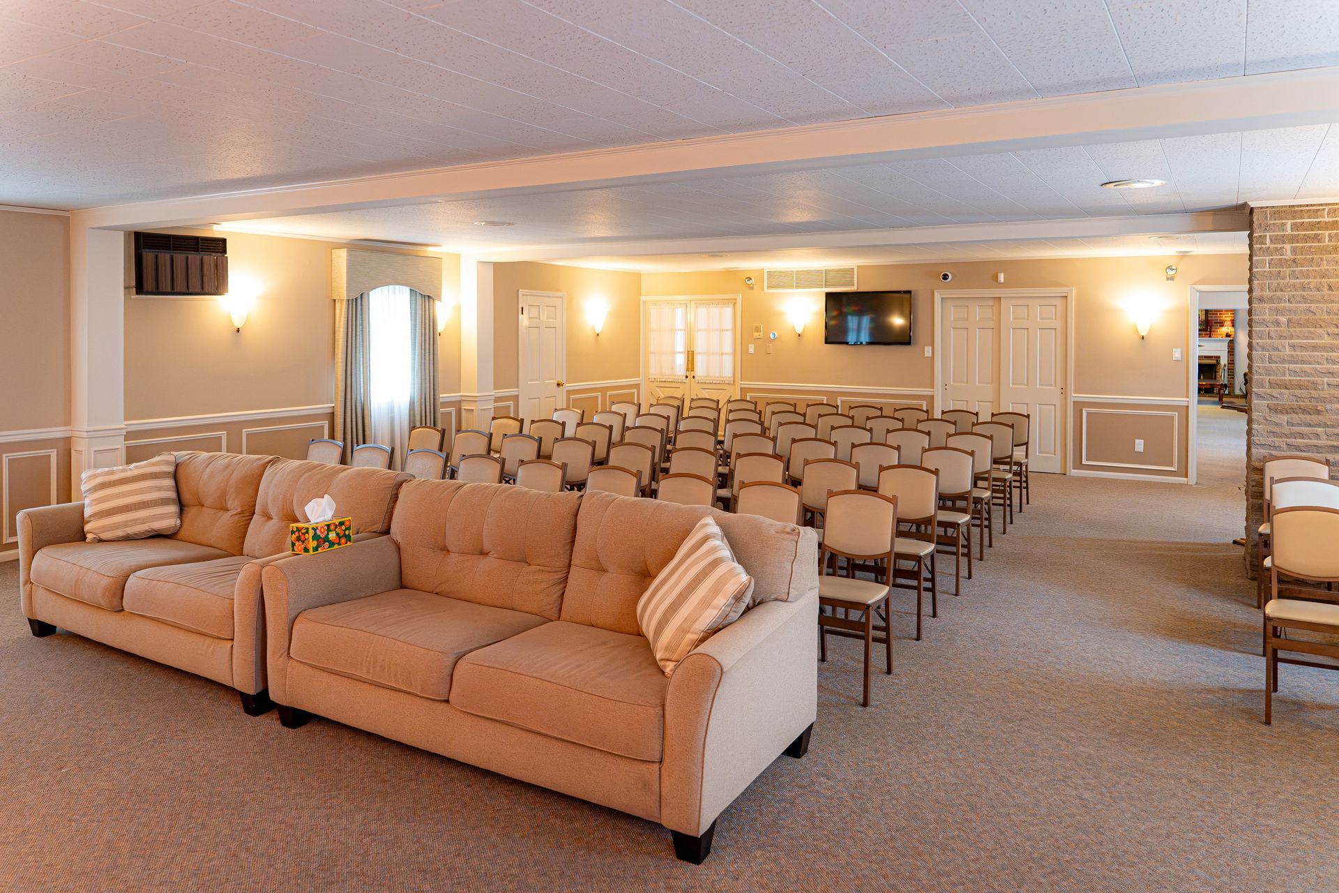 Bright, spacious waiting room with tan sofas and rows of chairs facing a wall-mounted TV
