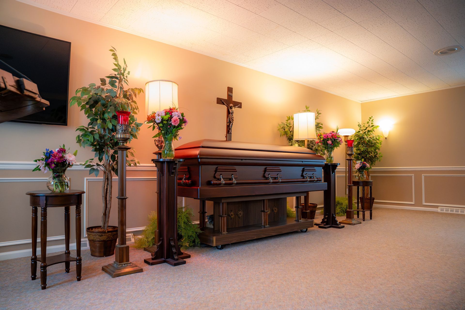 Memorial display with casket, flowers, lamps, and cross in a softly lit room