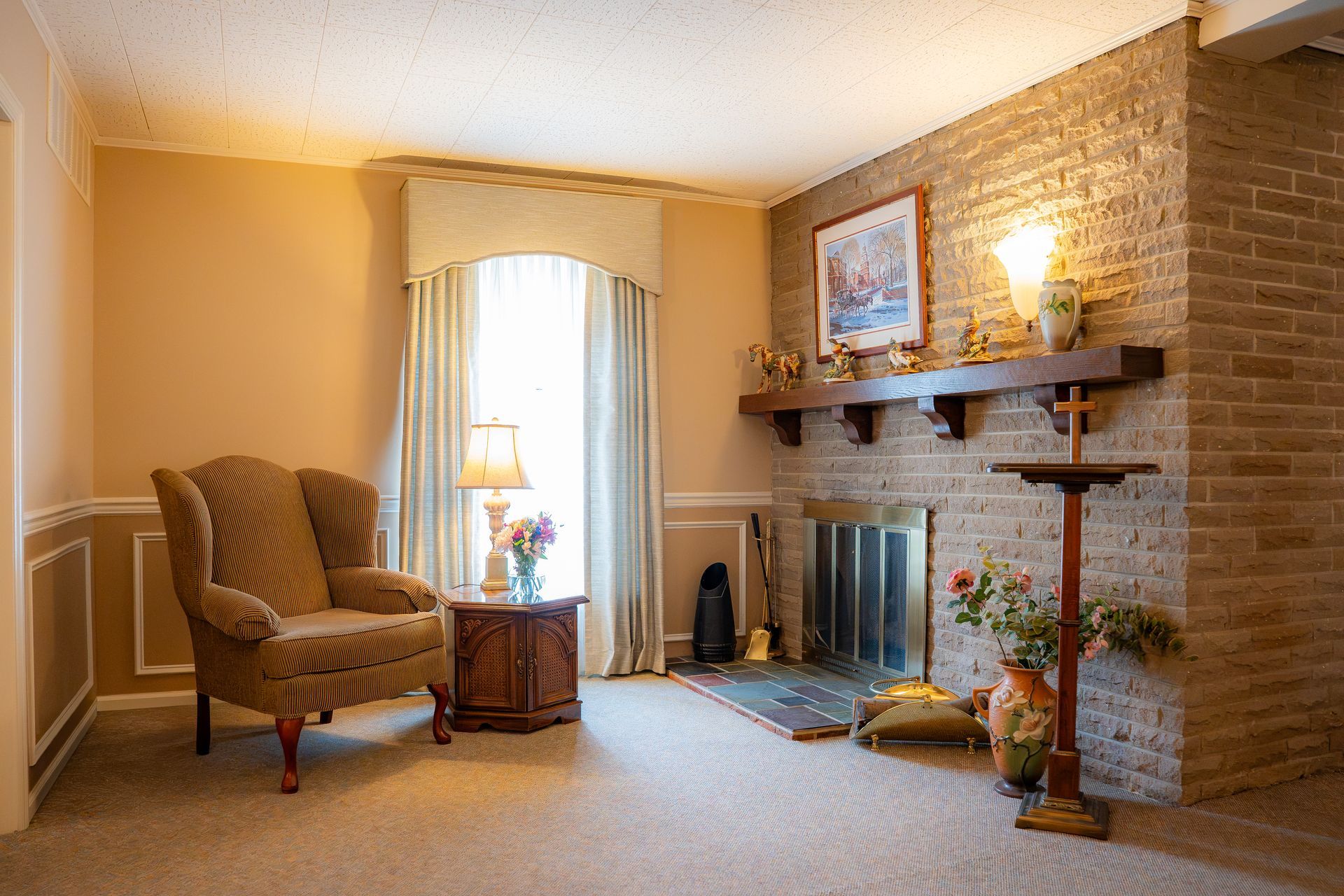 Cozy living room with armchair, fireplace, brick wall, and sunlit window with curtains.