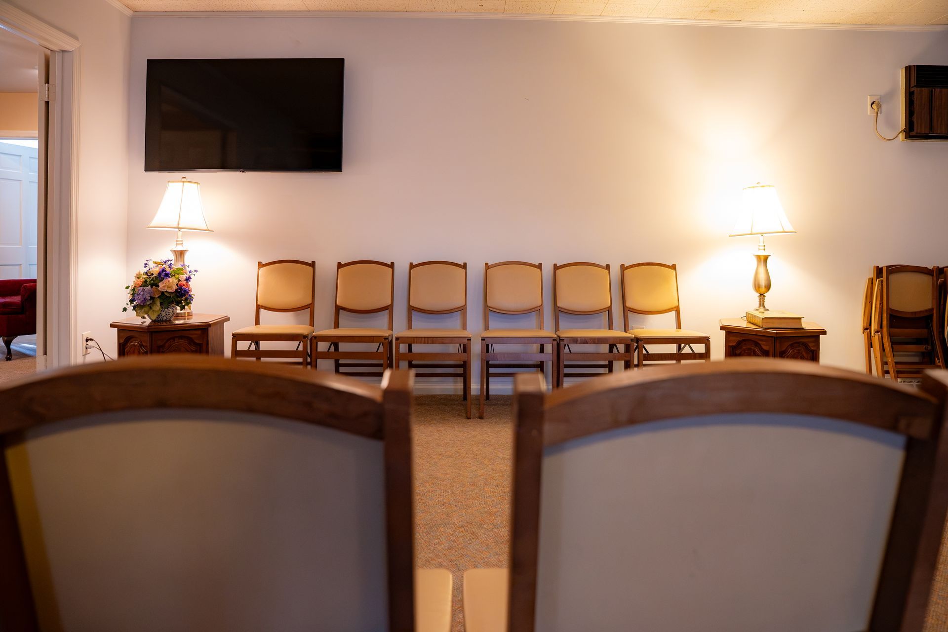 Empty meeting room with chairs, front table, wall-mounted TV, and warm lamps on either side.