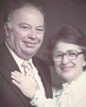 Smiling couple posing together in formal attire, with the woman’s hand on the man’s chest.