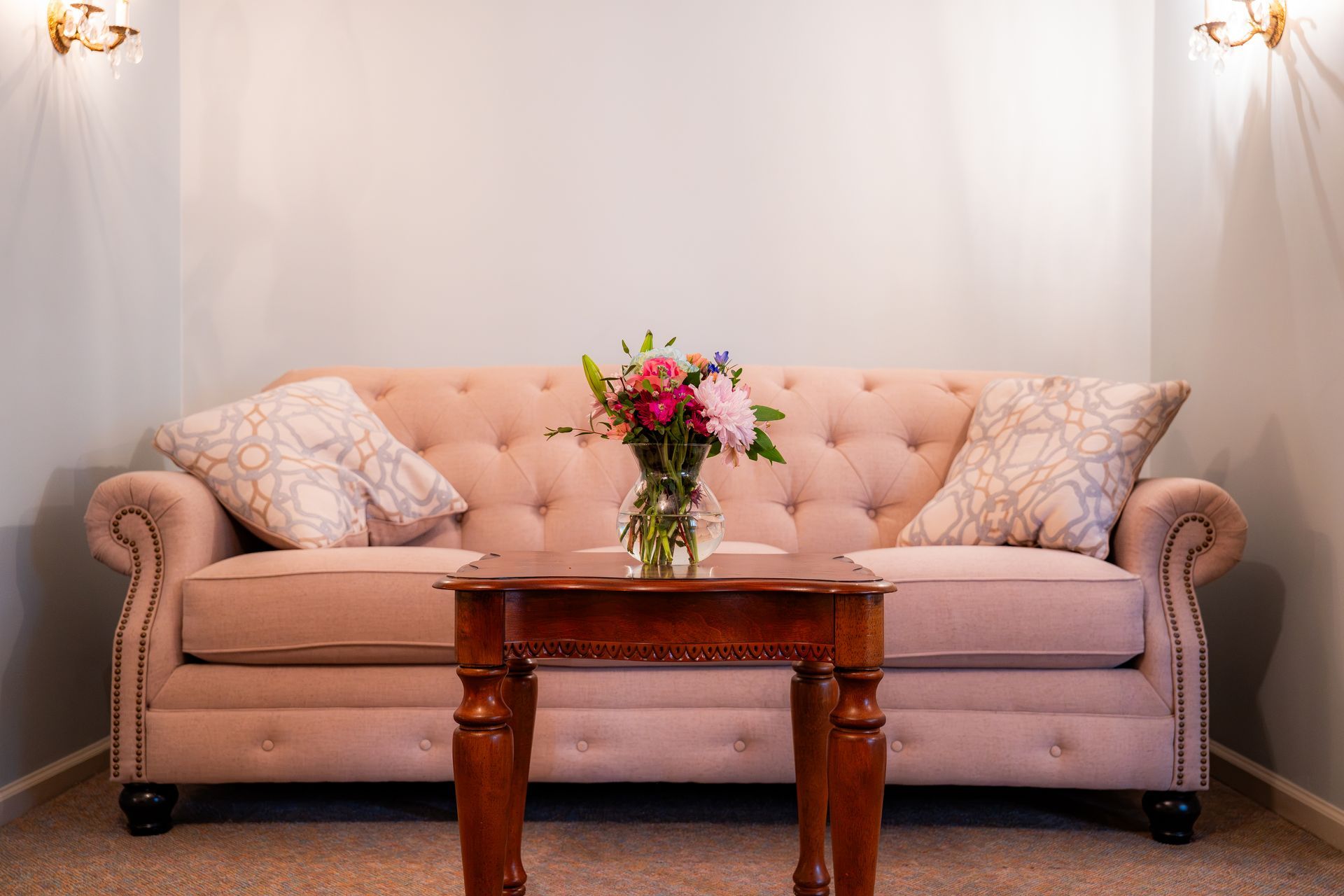 Pink tufted sofa with floral pillows and a vase of flowers in a softly lit room