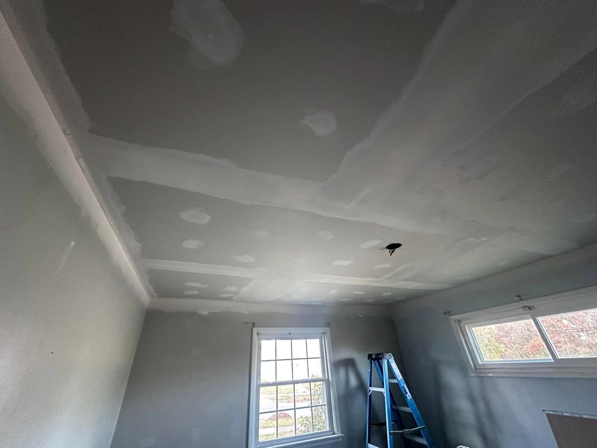Small room with unfinished drywall ceiling, ladder, window, and blue rolling scaffolding.