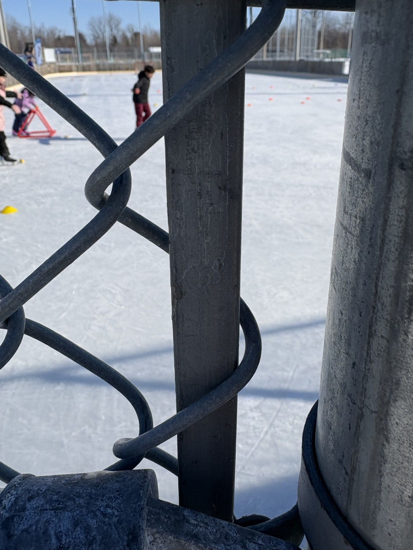 Clôture en grillage au premier plan ; personnes jouant au hockey sur glace à l'arrière-plan.