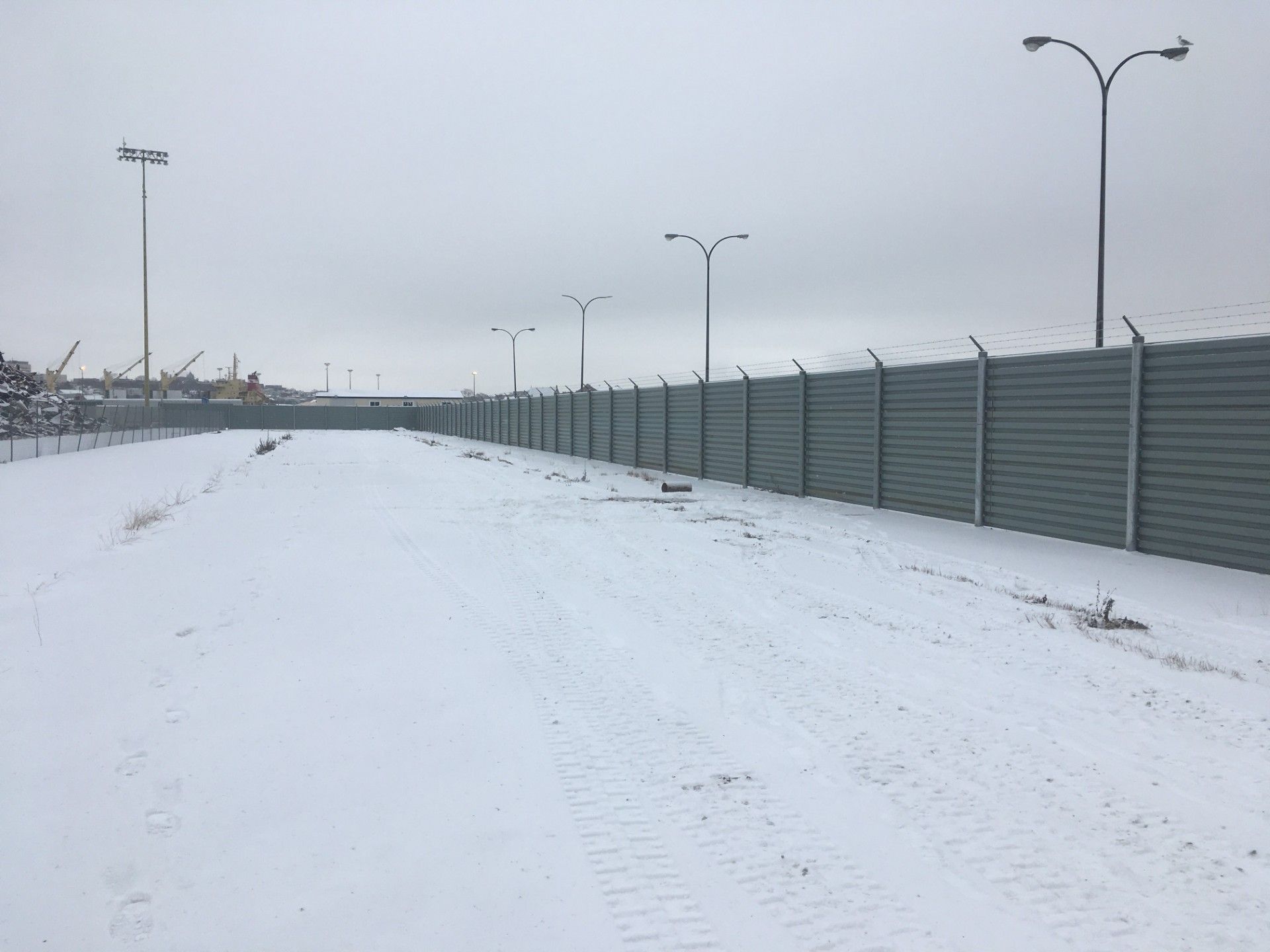 Snow-covered industrial area with a tall fence topped with barbed wire, and streetlights under a cloudy sky.