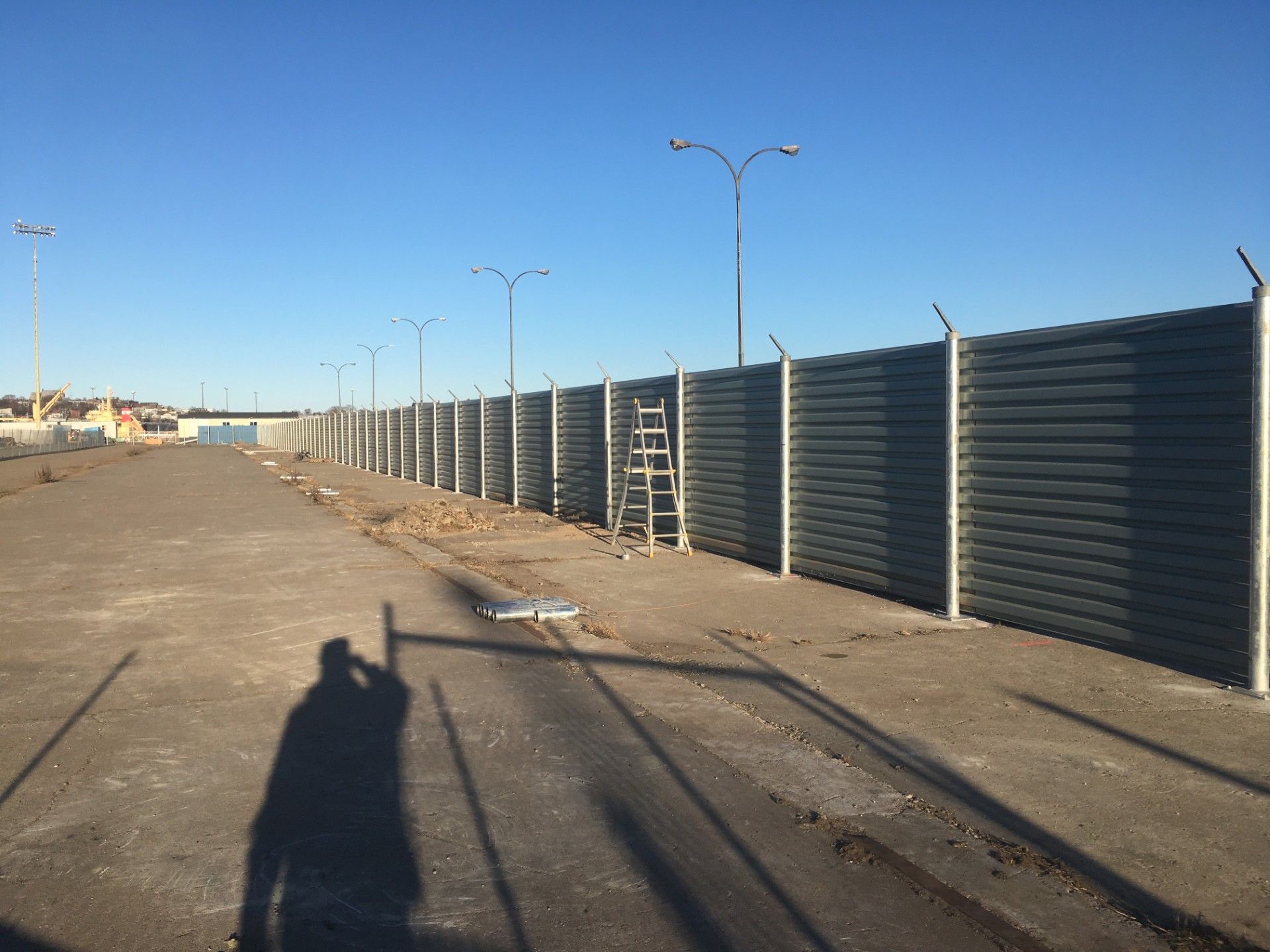 Long gray fence with barbed wire on top, next to an empty lot. A ladder leans against the fence.