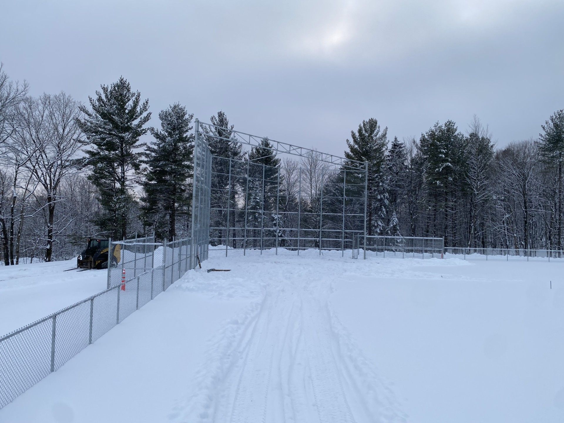 Snowy baseball field with chain-link fence, trees, and cloudy sky.