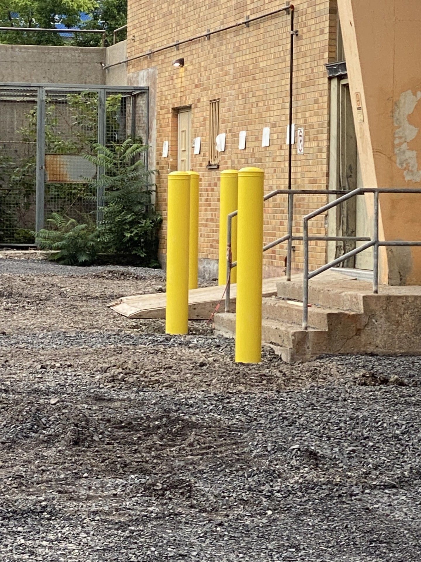 Yellow bollards in front of a building with a stairway and a gravelly ground surface.