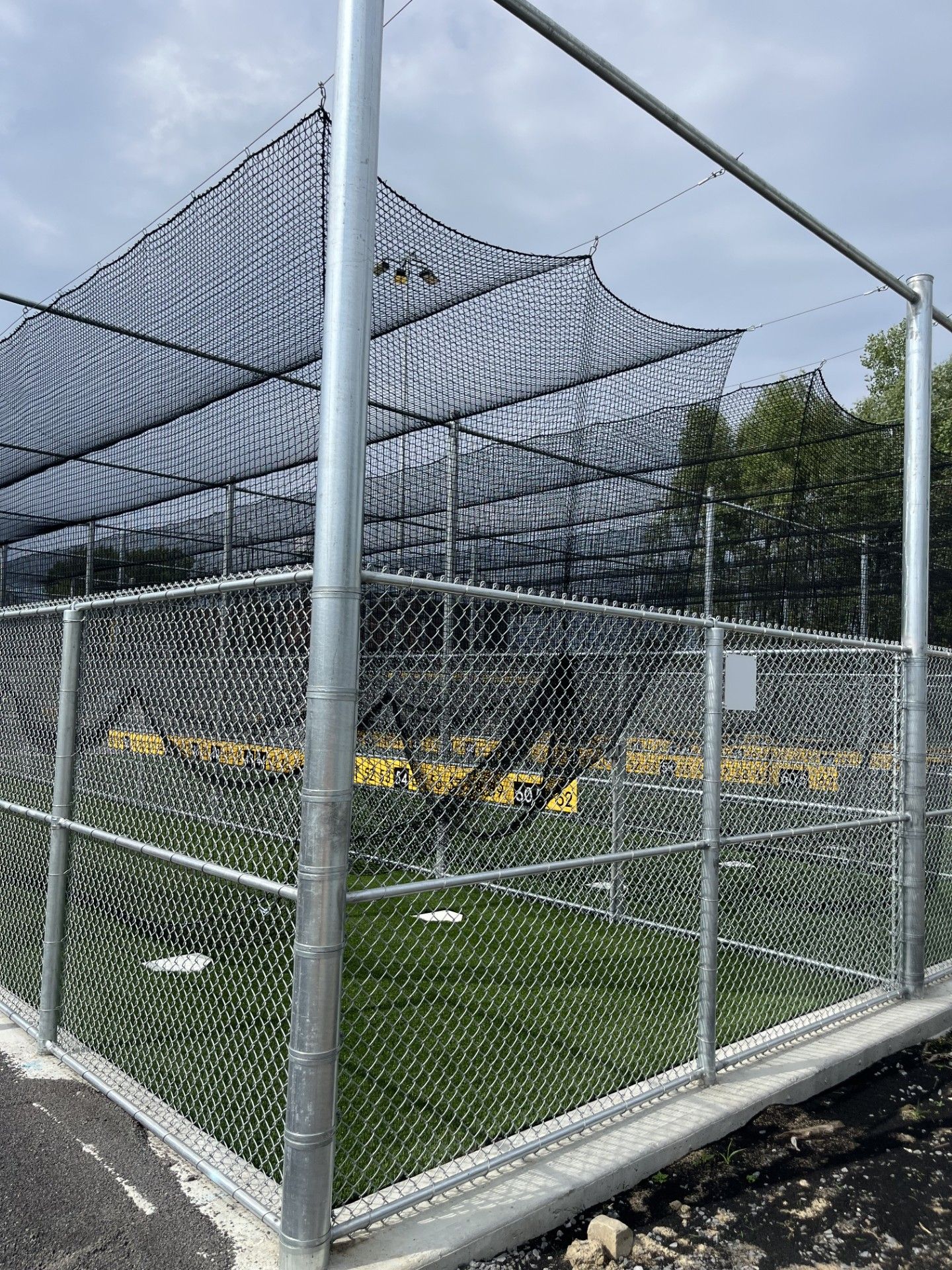Baseball batting cage enclosed by chain-link fence and netting. Green artificial turf inside. Gray metal poles.