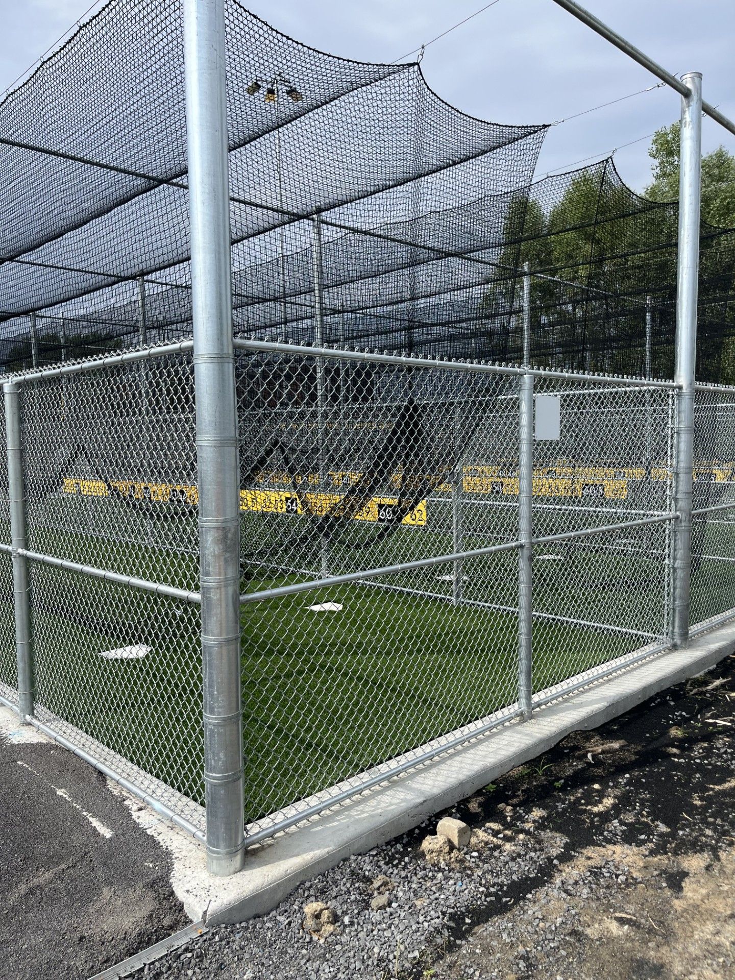 Baseball batting cage with chain-link fence, green turf, black netting, and concrete base.