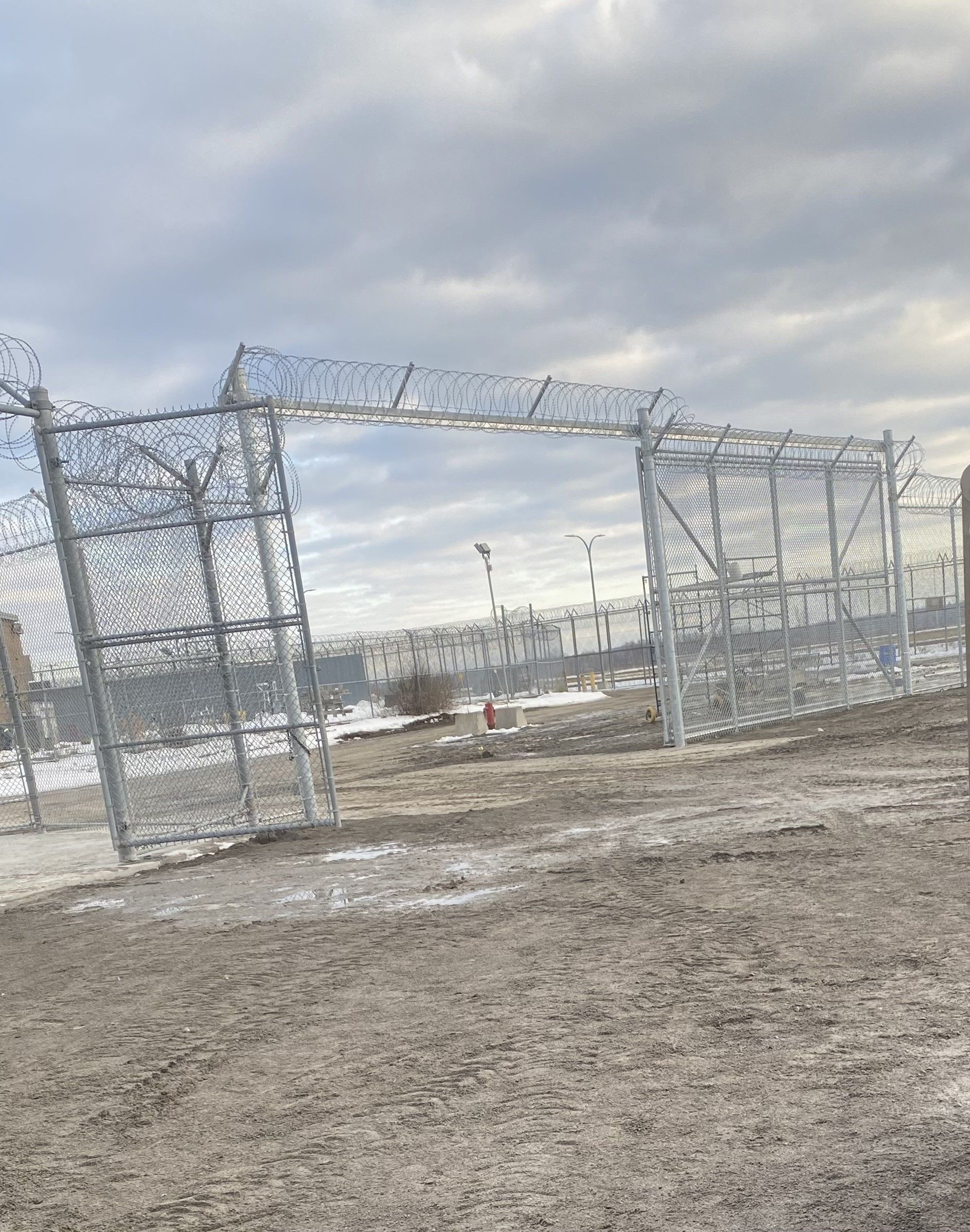 Open security gate with barbed wire atop a chain-link fence, set against a cloudy sky.