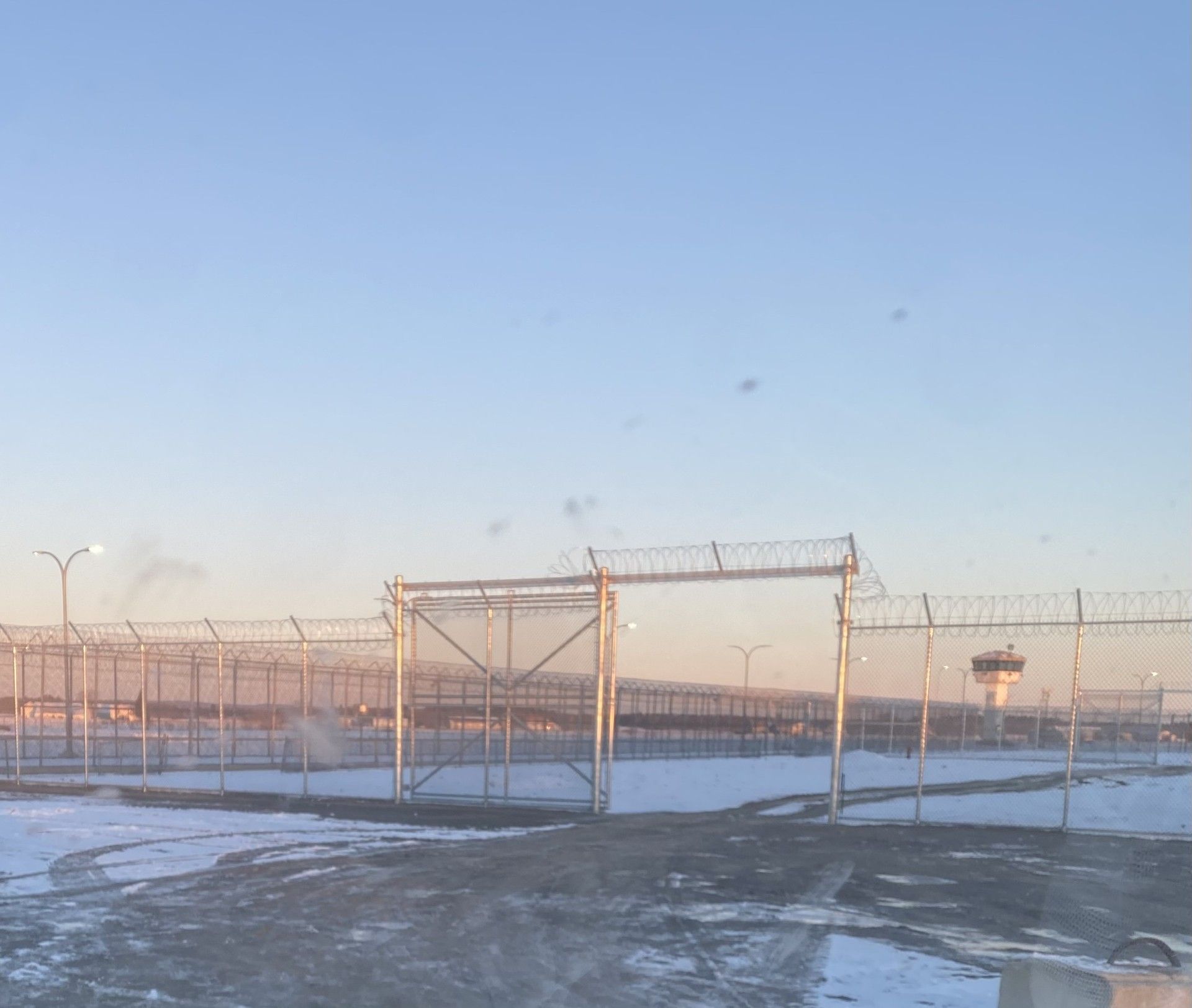 Fenced enclosure with barbed wire, snow-covered ground, and a control tower under a blue sky.