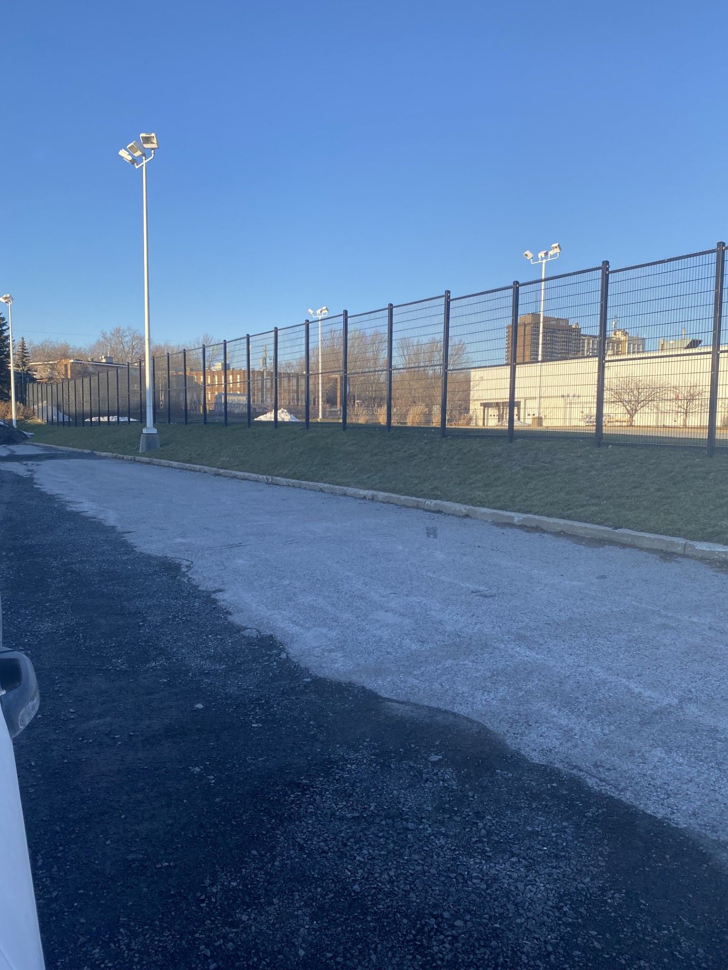 Paved path leading to a fenced area with light poles against a blue sky.