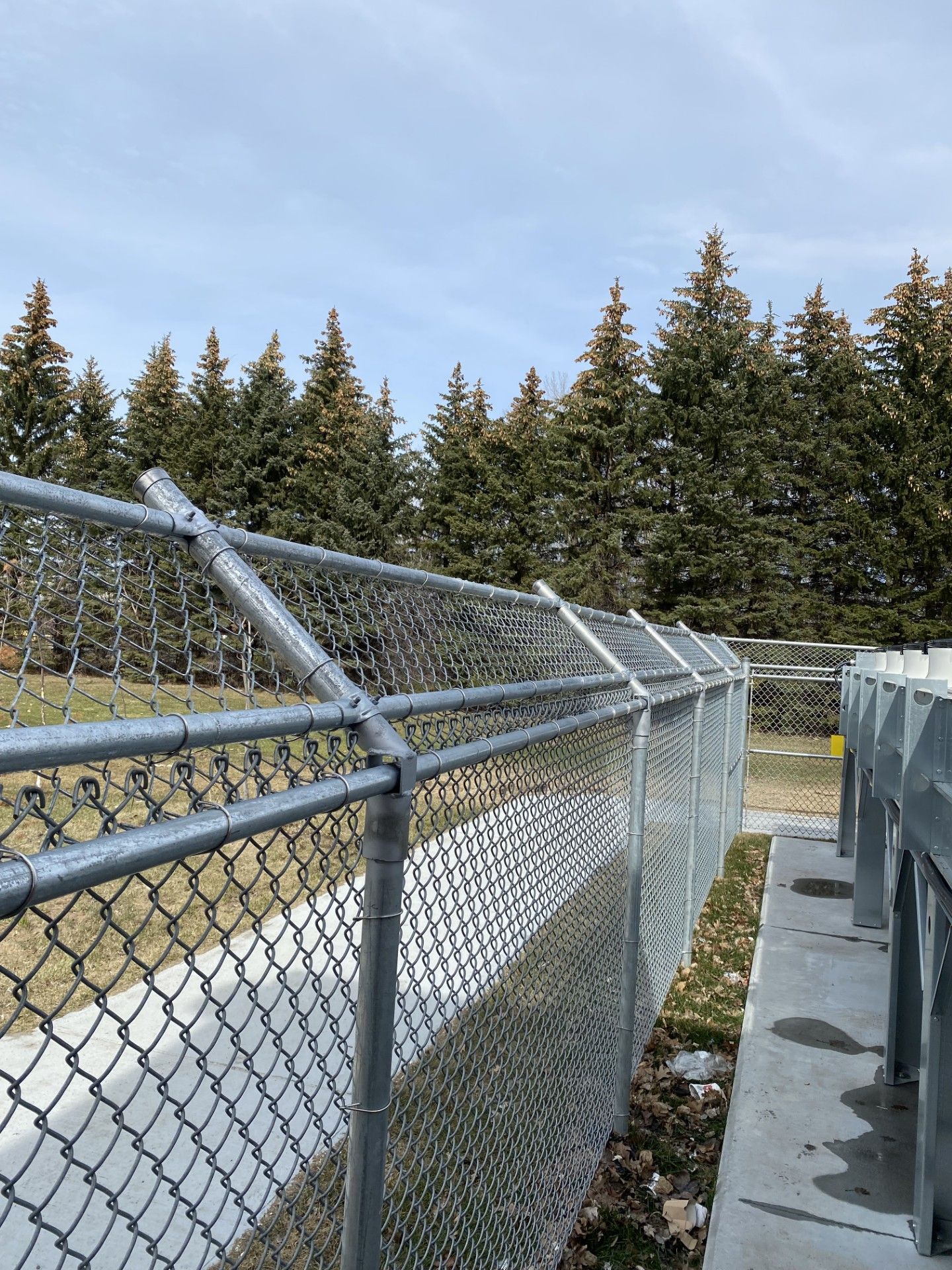 Chain-link fence with angled top, trees in the background, cloudy sky.