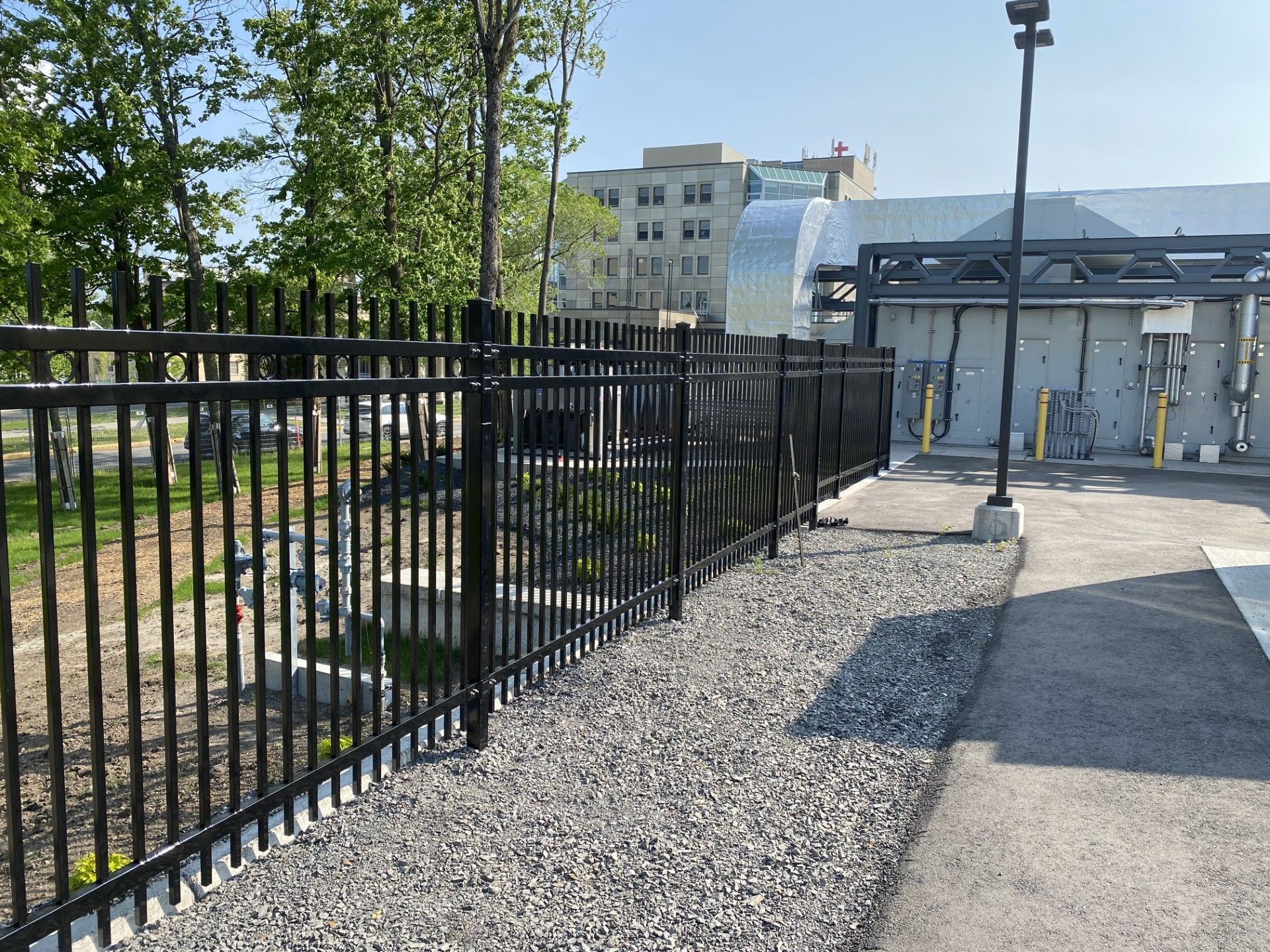 Black metal fence along a gravel path, next to a building with trees and the sky in the background.