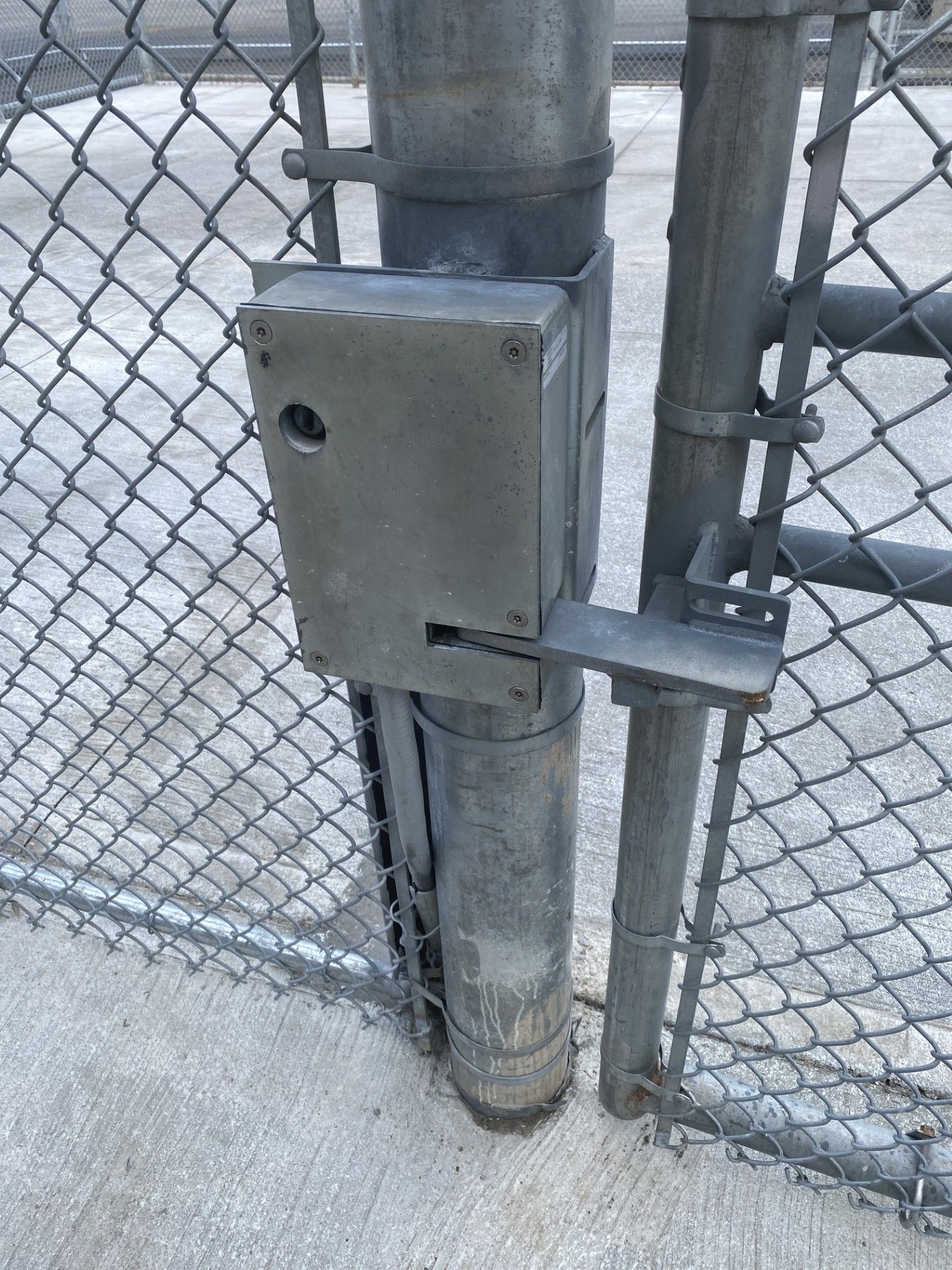 A metal gate latch and control box mounted on a chain link fence post.