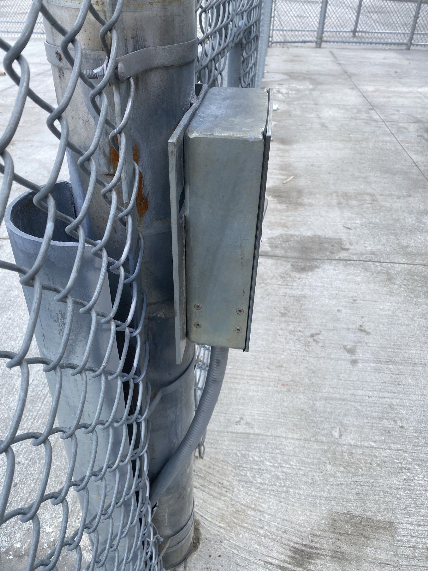 Metal electrical box attached to a chain-link fence post. Gray concrete background.