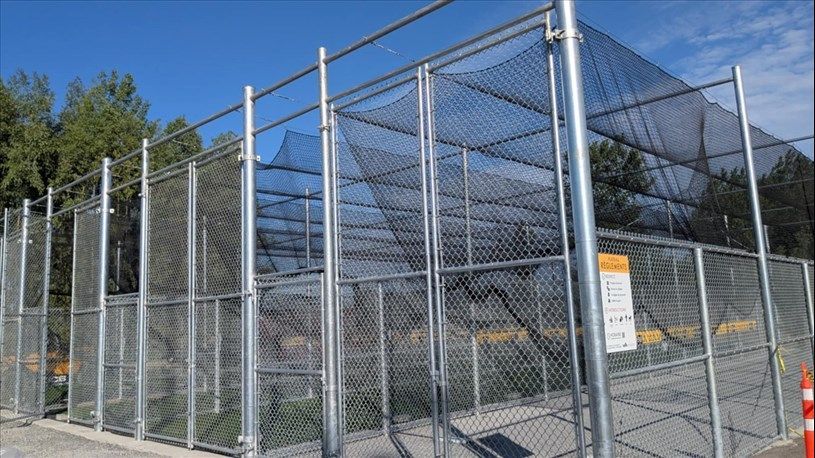 Chain-link fence enclosure with netting, on concrete, against a blue sky, possibly a batting cage.