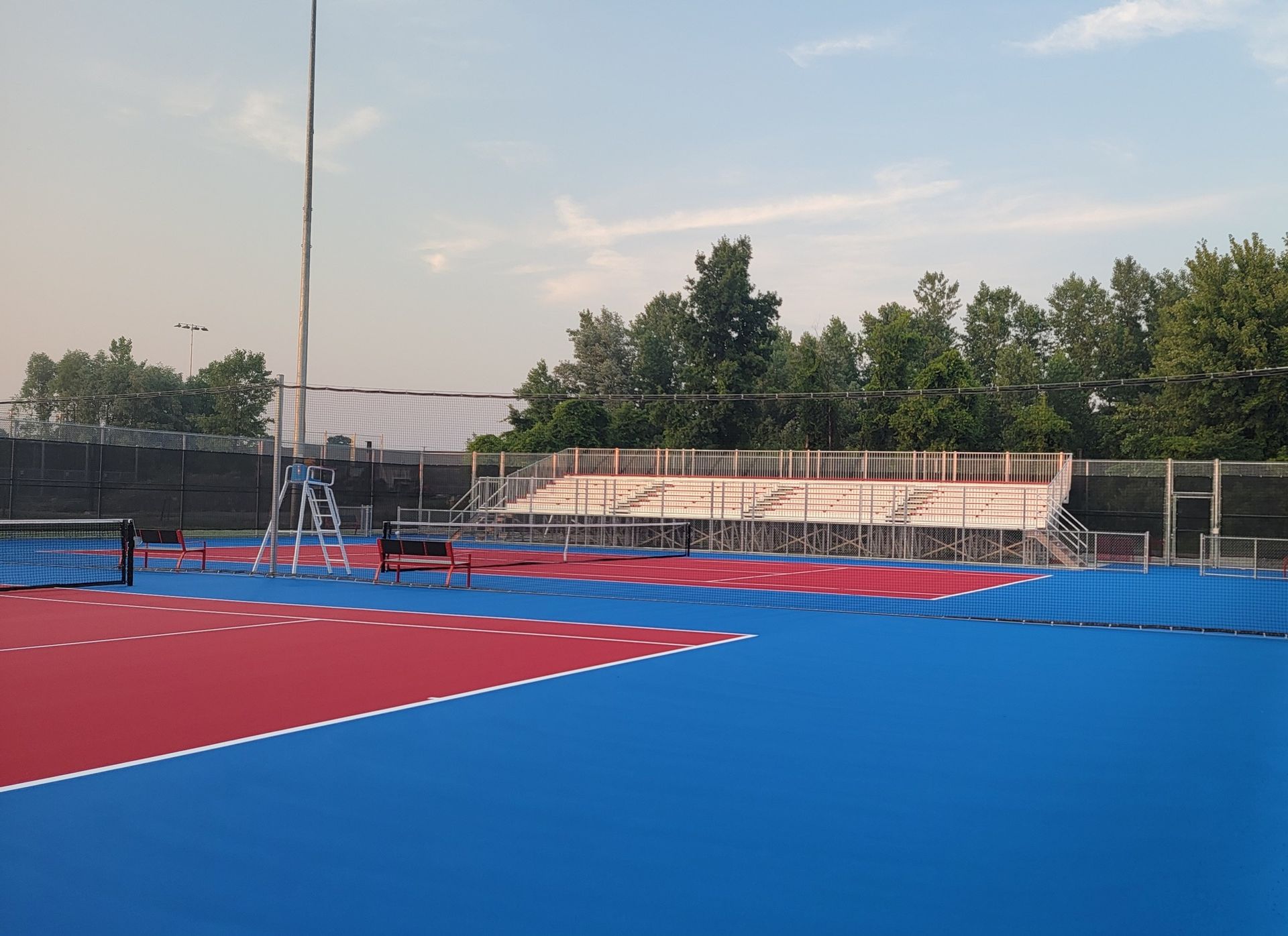 Blue and red sports court with bleachers, trees, and a tall pole against a blue sky.