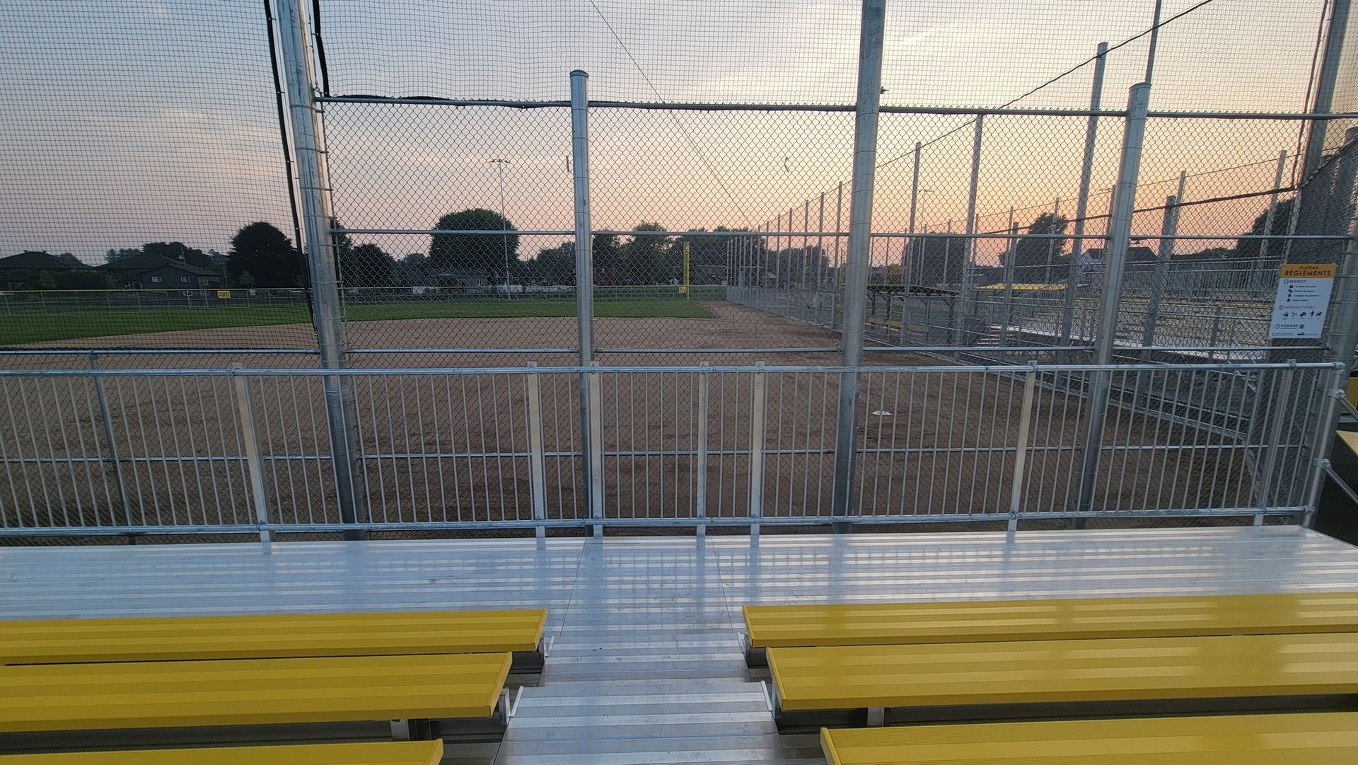 Yellow bleachers facing a baseball field, with a chain-link fence and dusk sky in the background.
