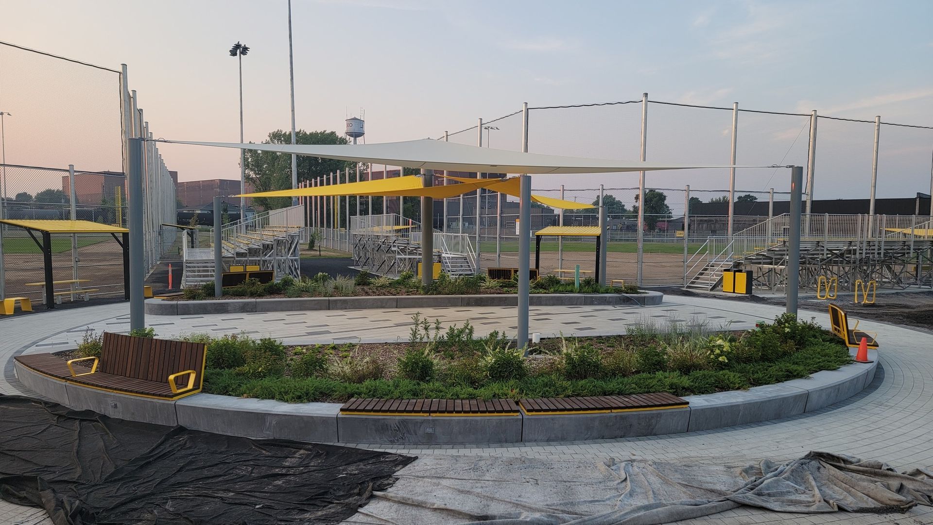 Circular park with benches, yellow canopies, plants, and netting. Construction in progress.