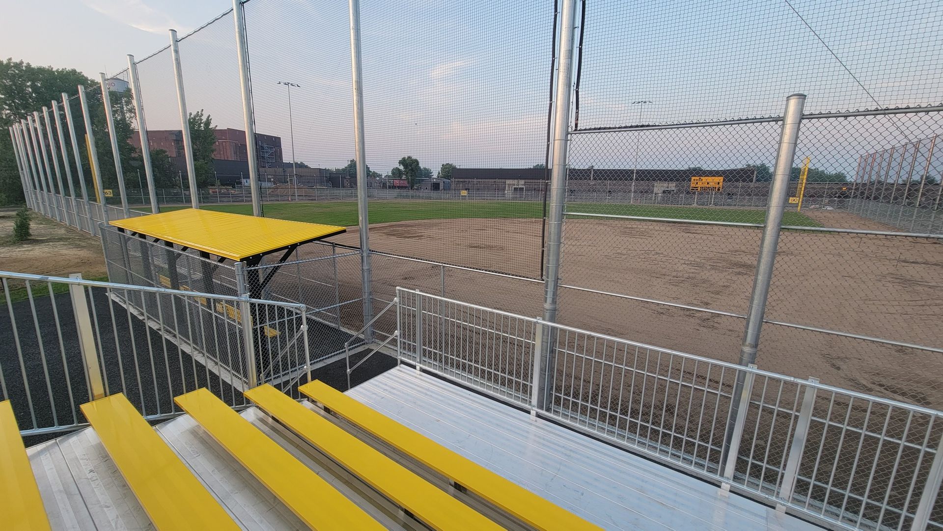 Bleachers overlook a sand volleyball court, enclosed by netting. Yellow benches, sunny day.