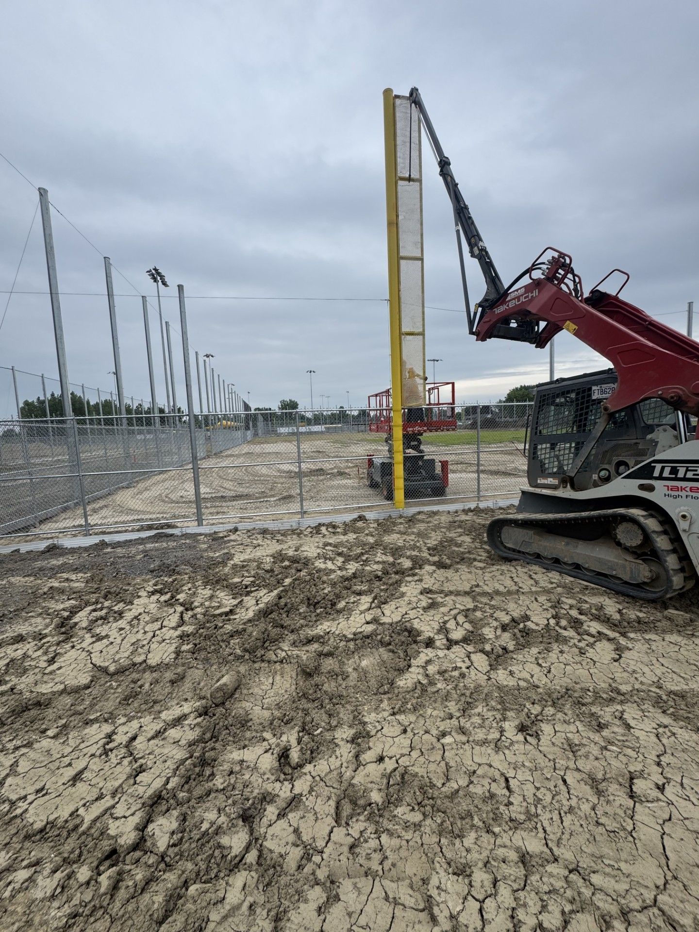 Construction of baseball field fence; skid steer with crane arm placing a yellow pole on muddy ground.
