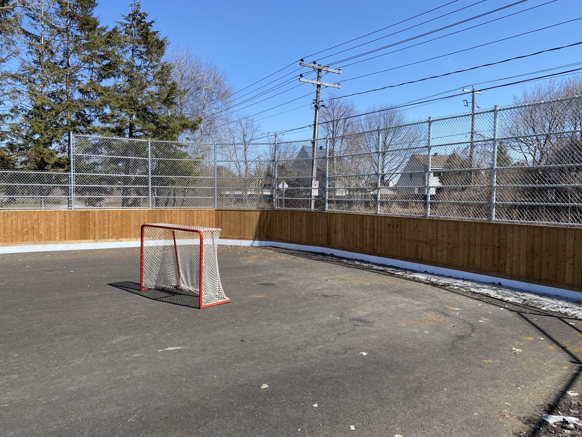 An empty hockey rink with a net, enclosed by a wooden barrier and chain-link fence, under a clear sky.