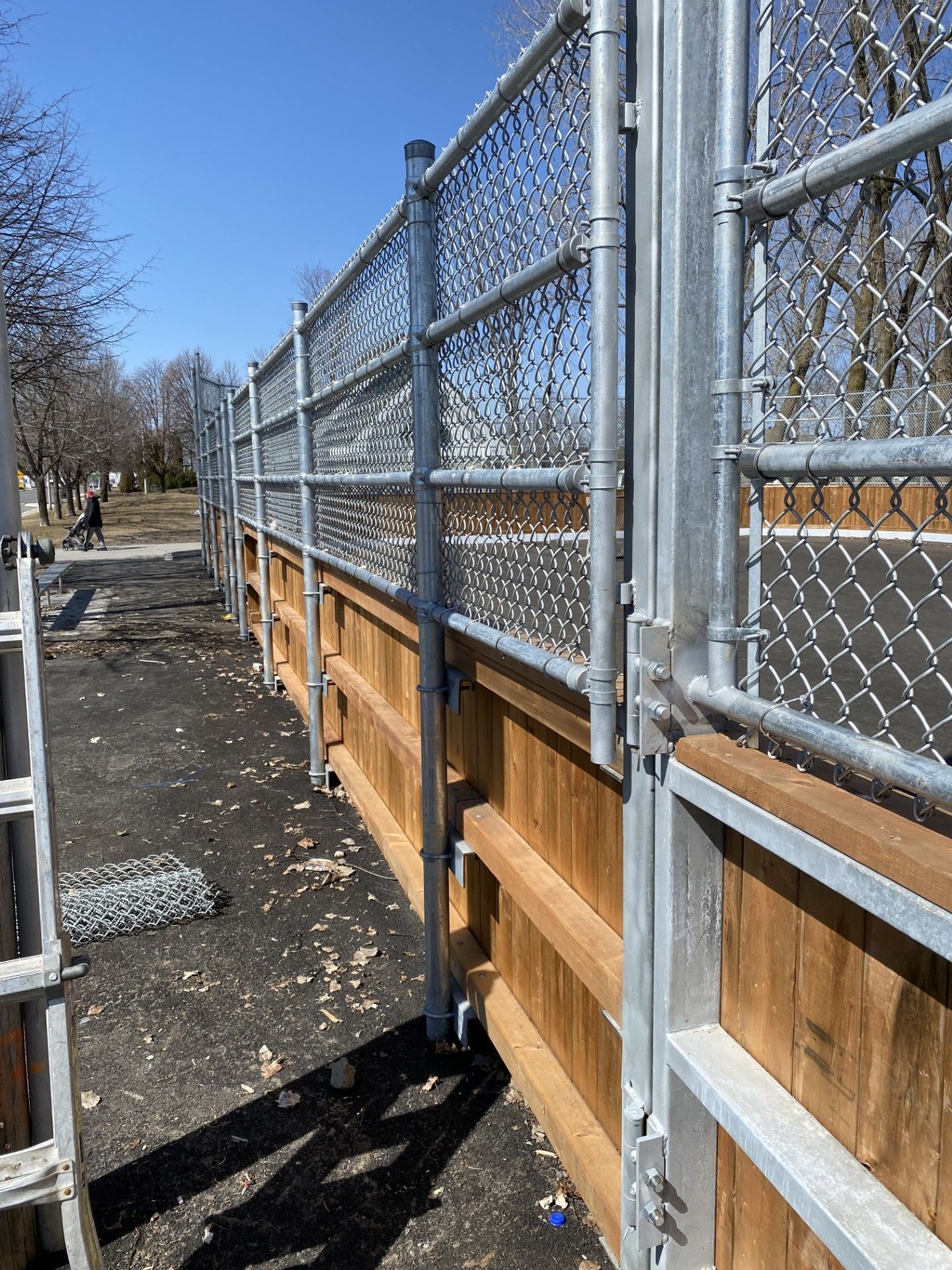 Chain-link fence atop a wooden privacy fence, beside a paved area under a blue sky.