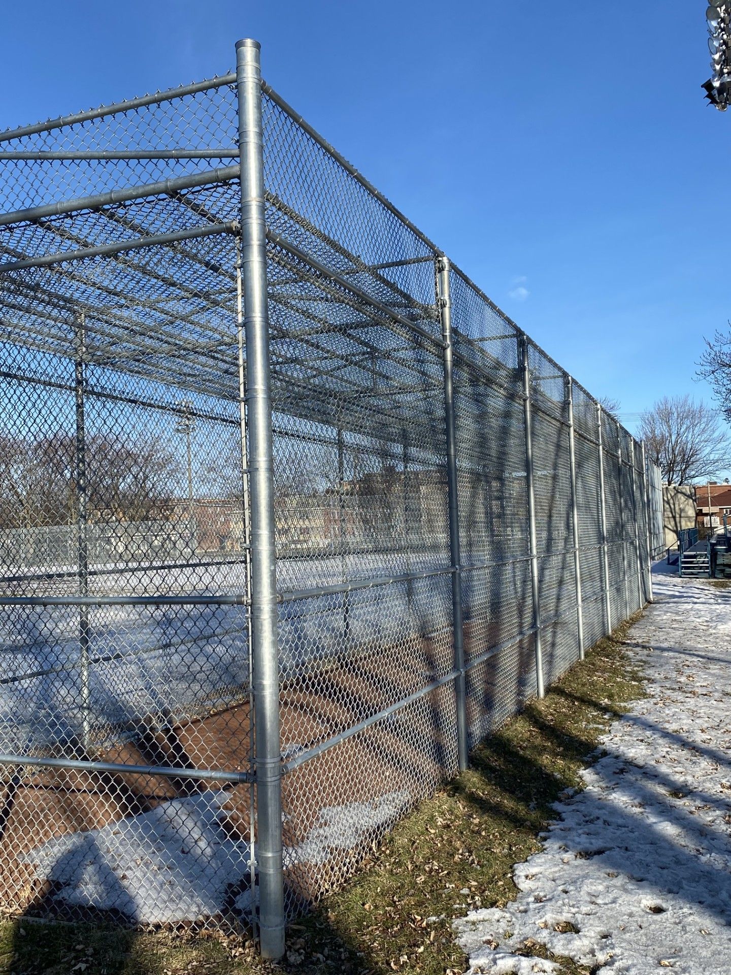 Chain-link fence surrounding a field with patches of snow and a clear, blue sky.