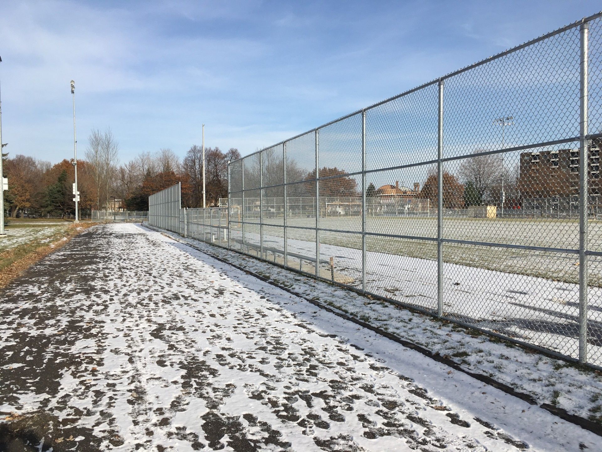 Snowy path next to a chain-link fence bordering a sports field on a sunny day.