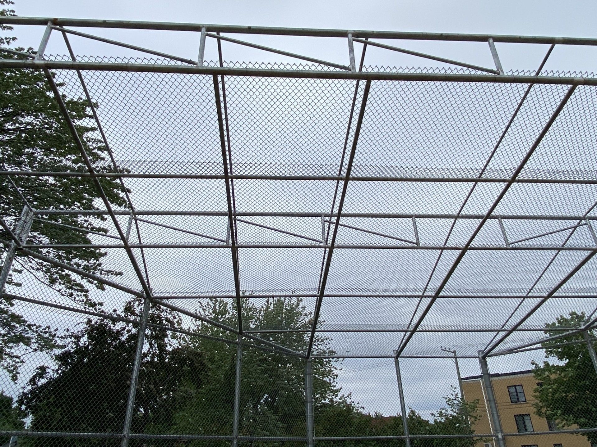 Metal cage with netting overhead, likely for sports, against a cloudy sky.