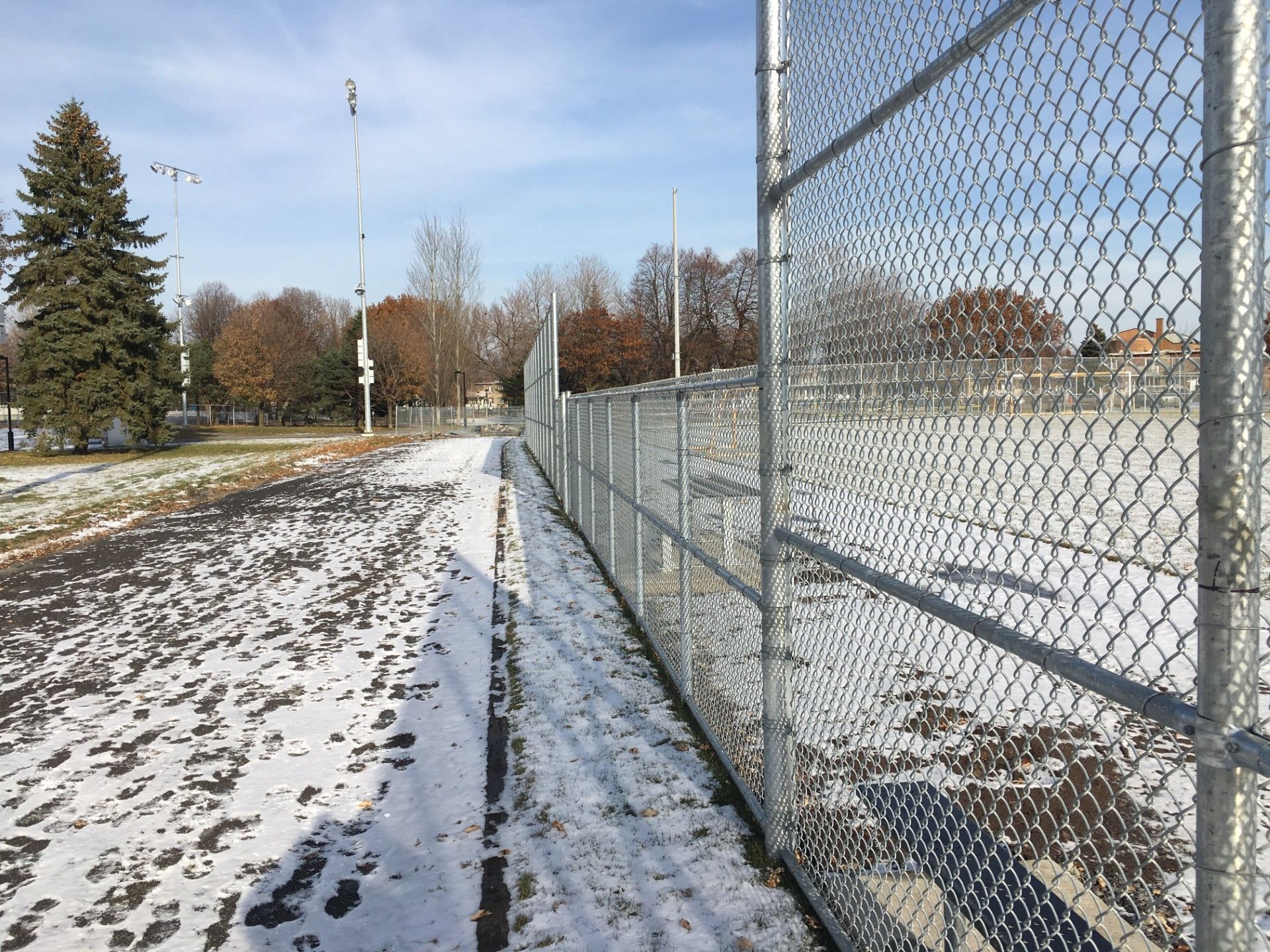 A snow-covered path next to a chain-link fence, trees, and a blue sky on a cold day.