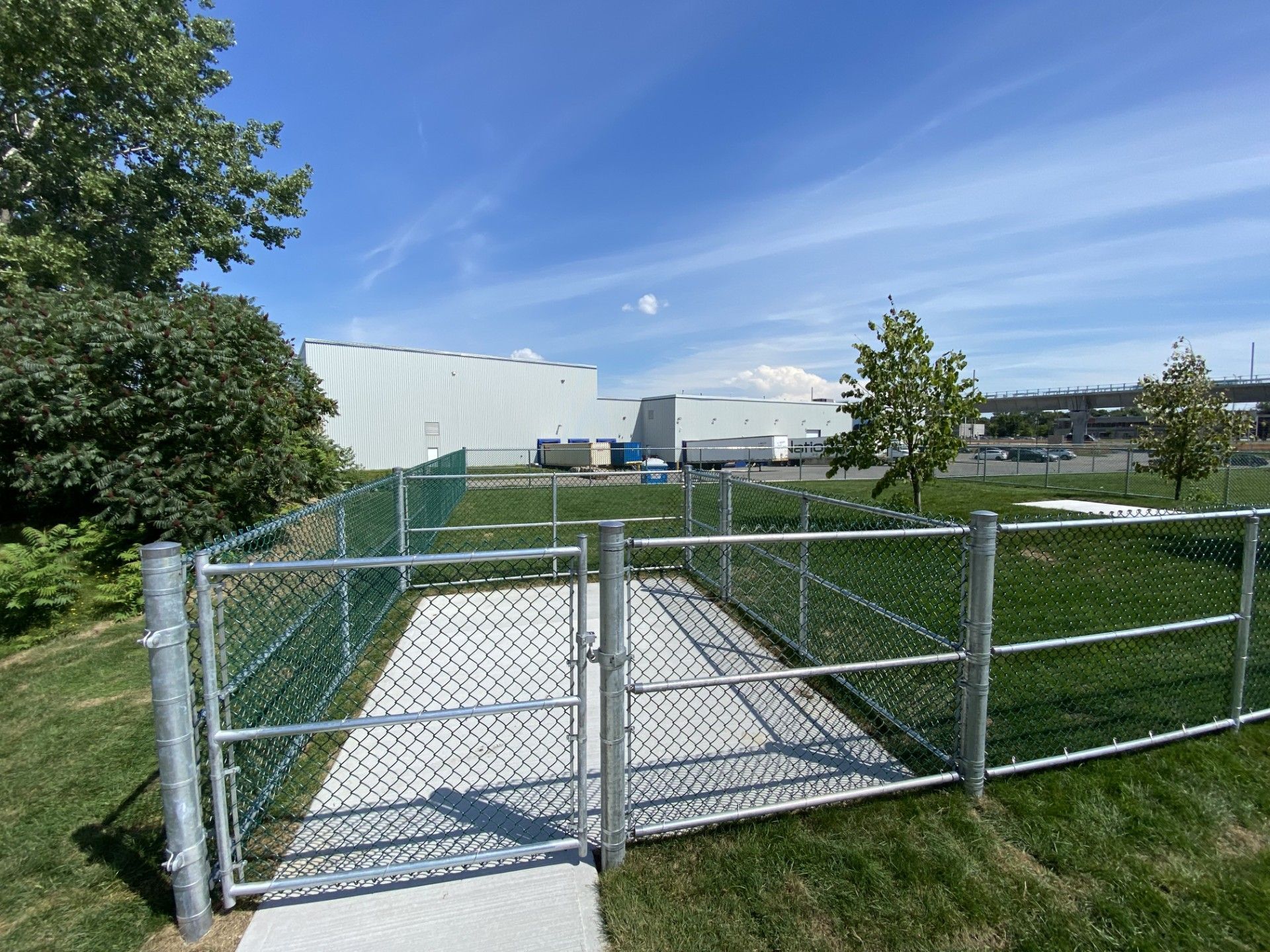 Fenced-in area with gate, concrete path. Green grass, blue sky, and industrial building in the background.